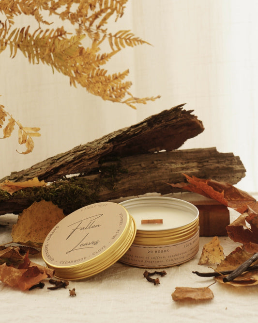 candle nest doftljus "Fallen leaves" i rund metall burk av trä står på ett vitt underlag. Locket står lutat mot kanten och exponerar vaxet och veken av trä. Ljuset är omgivet av nedfallna löv, kryddnejlika, vaniljstänger och i bakgrunden syns mörk ved.