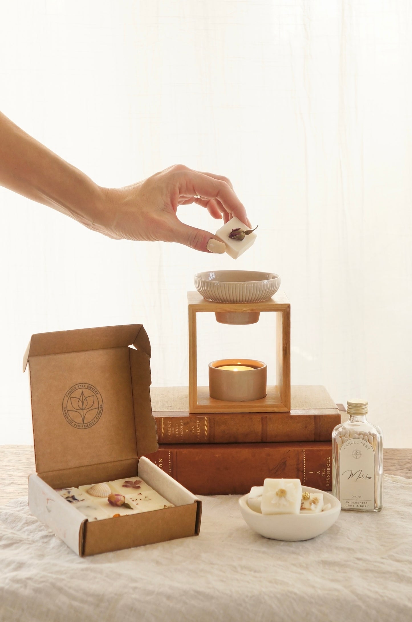 Hand som lägger en doftpralin i en aromalmapa. På bordet står en ask med doftpraliner, en flaska med tändstickor och en aromalampa i bambu och keramik.