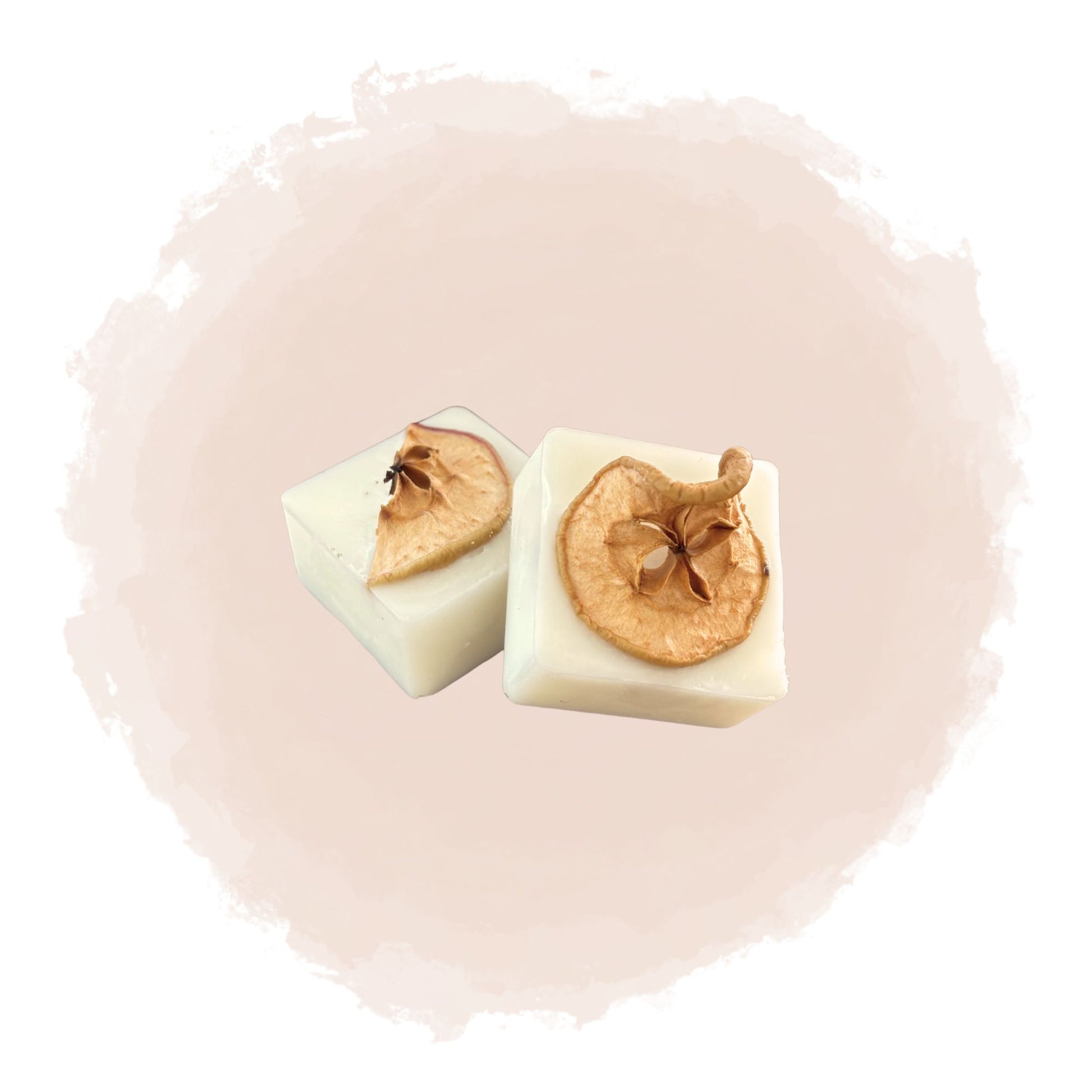 Två kubiska doftpraliner syns mot en beige bakgrund. Doftvaxen är dekorerade med skivor av torkat äpple