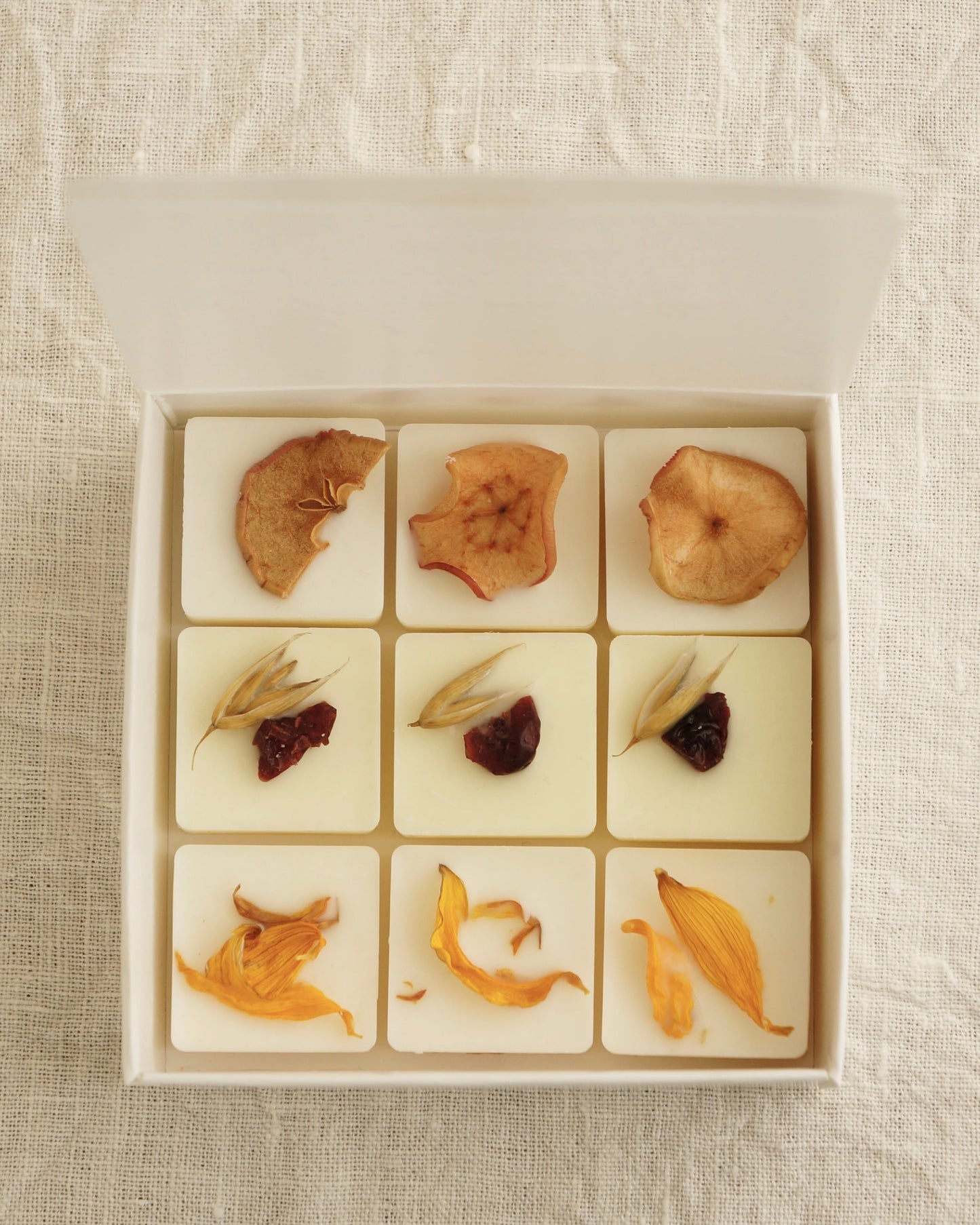 Candle nest ask med 9 doftpraliner (doftvax) står på en vit duk. De små kvadratisk vita vaxbitarna är dekorerade med torkade äpplen, nakenhavre, tranbär och torkade kronblad från solros. Bilden är tagen uppifrån. 