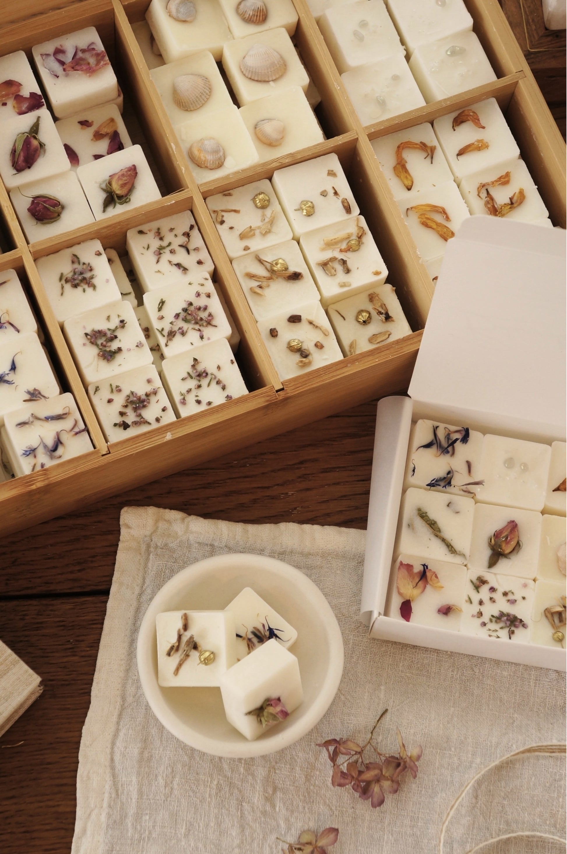 Bilden visar en telåda i bambu fylld med doftpraliner som är dekorerade med torkade blommor av olika slag. Lådan strå på ett bord av mörkt trä. På bilden syns även en ask med doftpraliner och en skål för doftpraliner där tre kubiska doftpraliner ligger i skålen. 