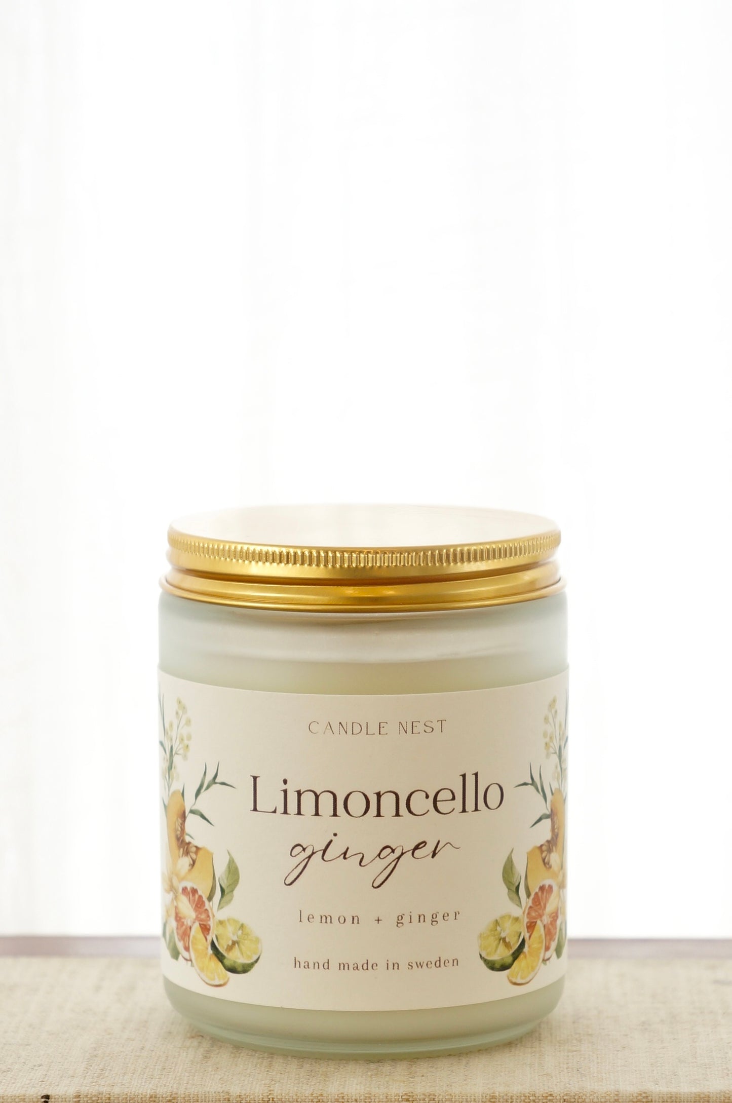 Ett doftljus i en frostad vit glasburk står på en bok. Runt ljuset finns ett off-white papper med bilder av frukter och texten "Limoncello ginger". Ljuset har ett skruvlock i guld lutat.