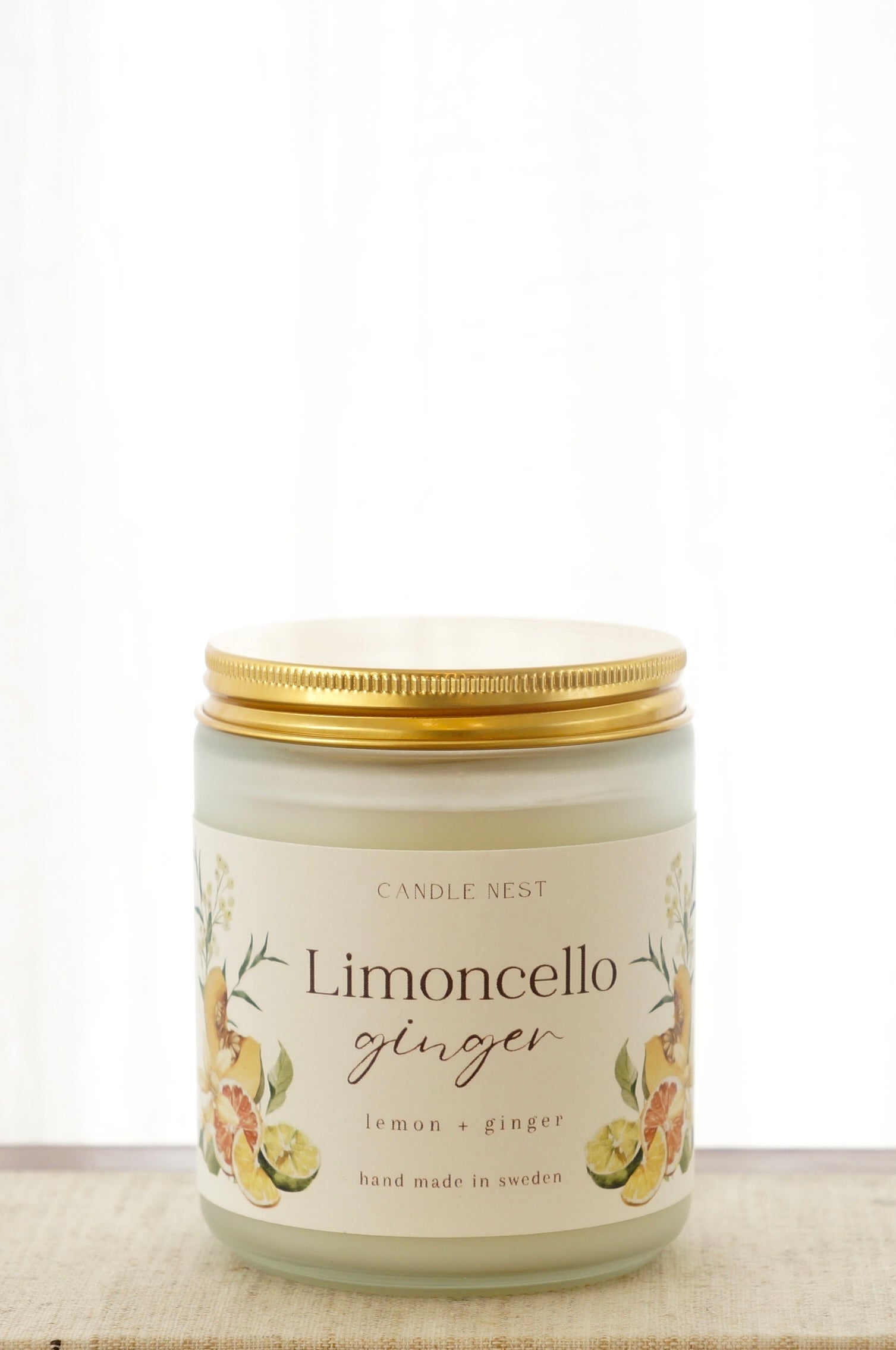 Ett doftljus i en frostad vit glasburk står på en bok. Runt ljuset finns ett off-white papper med bilder av frukter och texten "Limoncello ginger". Ljuset har ett skruvlock i guld lutat.