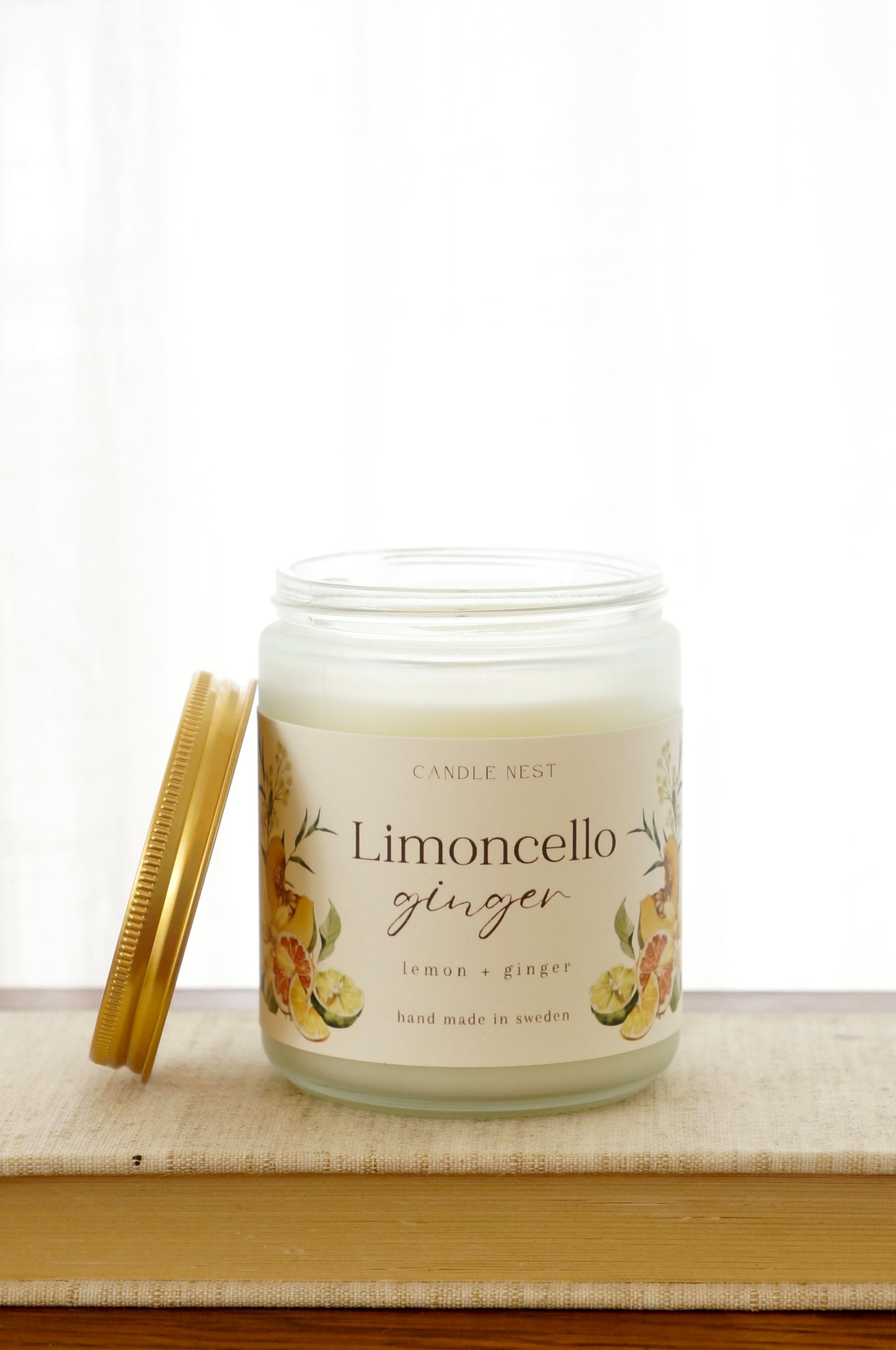 Ett doftljus i en frostad vit glasburk står på en bok. Runt ljuset finns ett off-white papper med bilder av frukter och texten "Limoncello ginger". Mot ljuset står burken skruvlock i guld lutat.