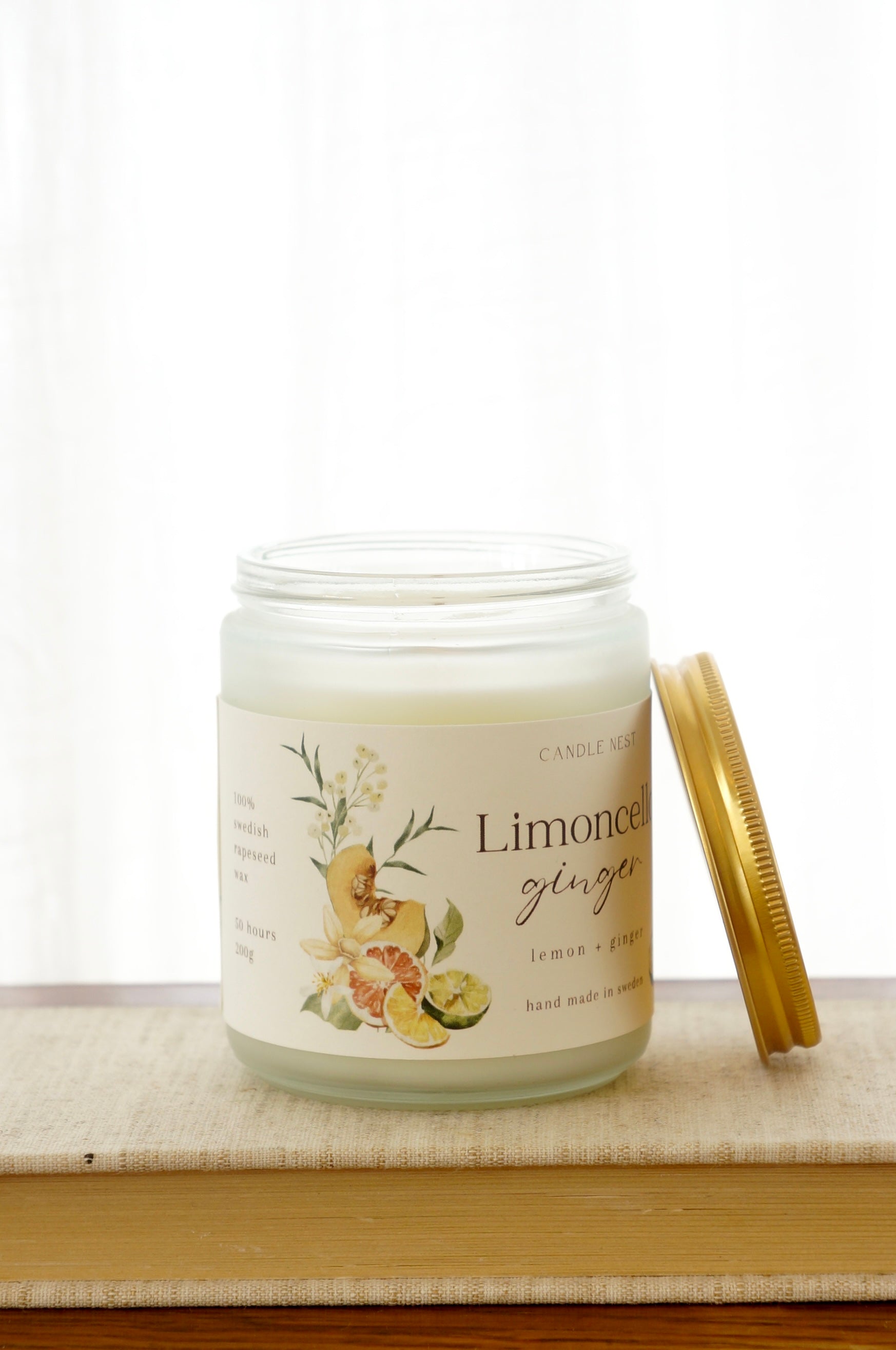 Ett doftljus i en frostad vit glasburk står på en bok. Runt ljuset finns ett off-white papper med bilder av frukter och texten "Limoncello ginger". Mot ljuset står burken skruvlock i guld lutat.