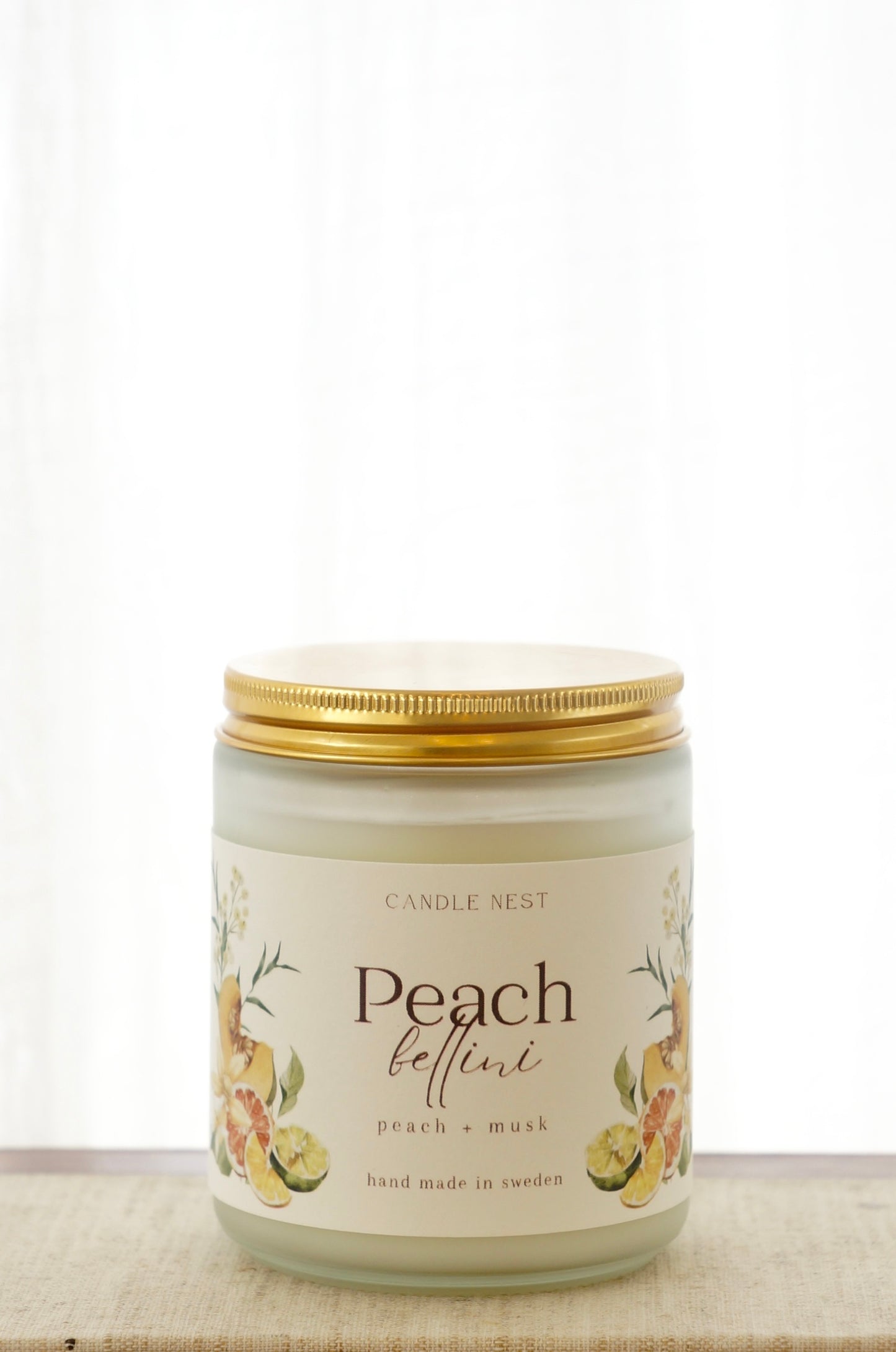 Ett doftljus i en frostad vit glasburk står på en bok. Runt ljuset finns ett off-white papper med bilder av frukter och texten "Peach bellini". Ljuset har ett skruvlock i guld.