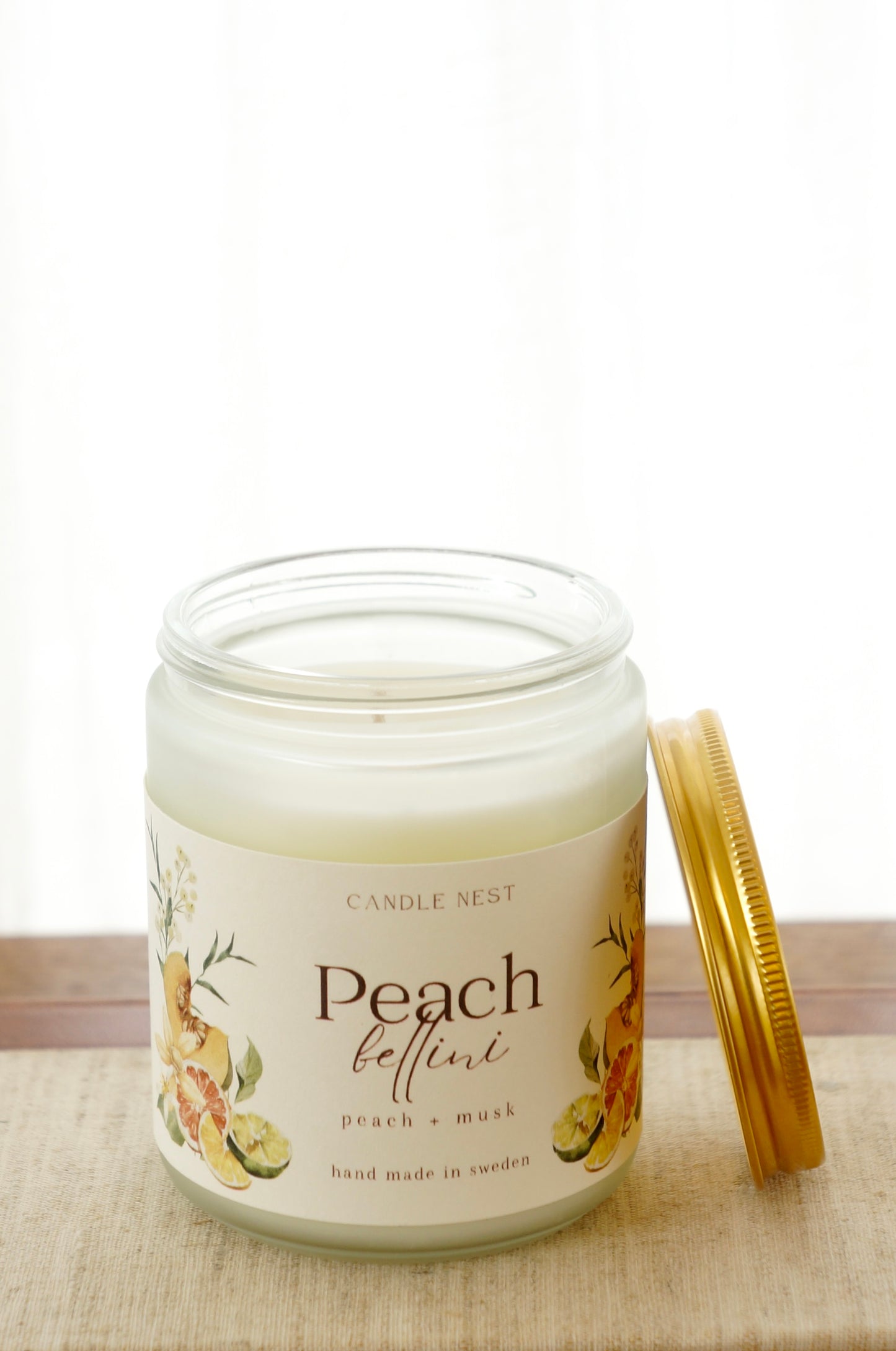 Ett doftljus i en frostad vit glasburk står på en bok. Runt ljuset finns ett off-white papper med bilder av frukter och texten "Peach bellini". Mot ljuset står burken skruvlock i guld lutat.