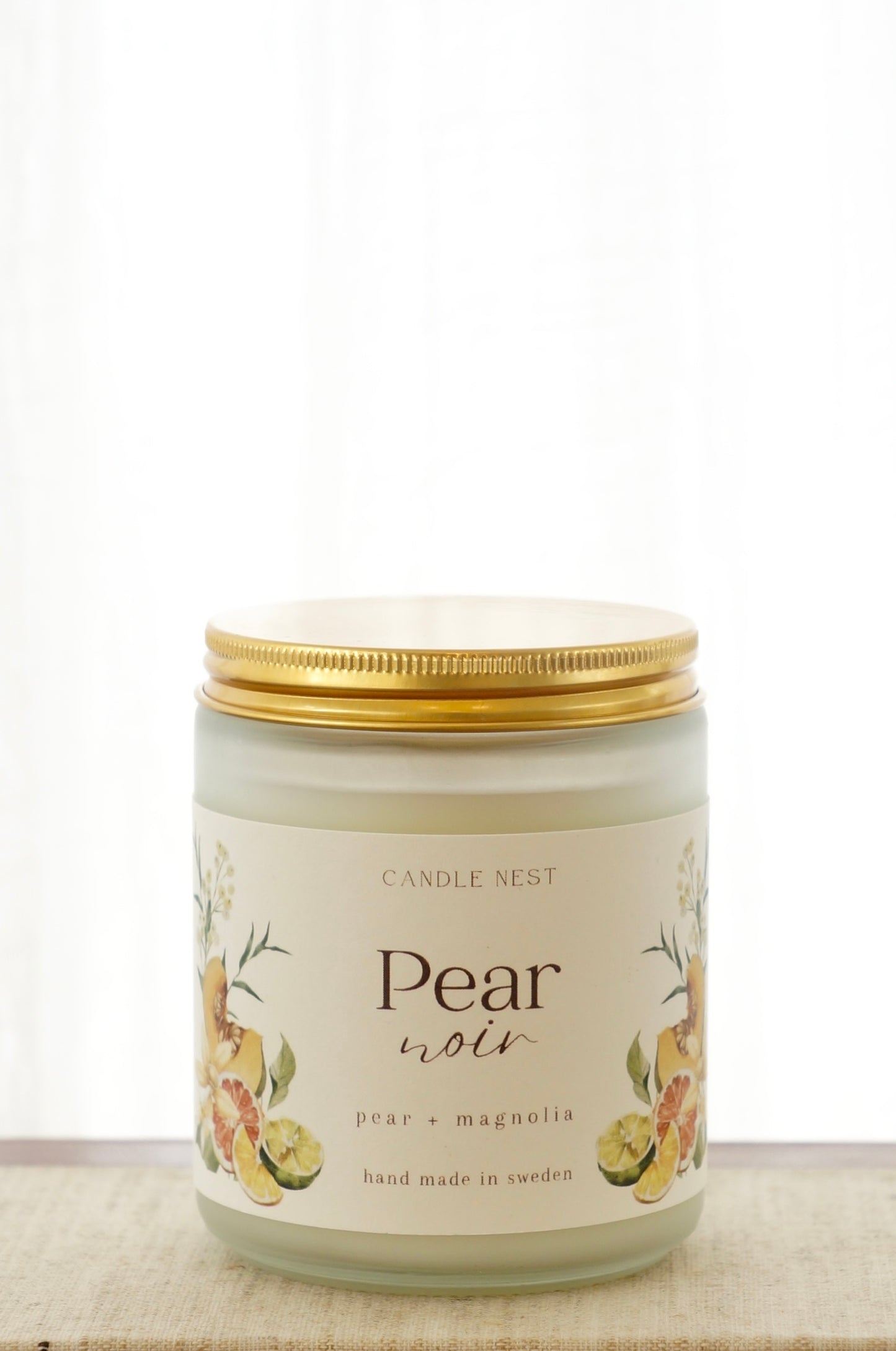 Ett doftljus i en frostad vit glasburk står på en bok. Runt ljuset finns ett off-white papper med bilder av frukter och texten "Pear noir". Ljuset har ett skruvlock i guld.