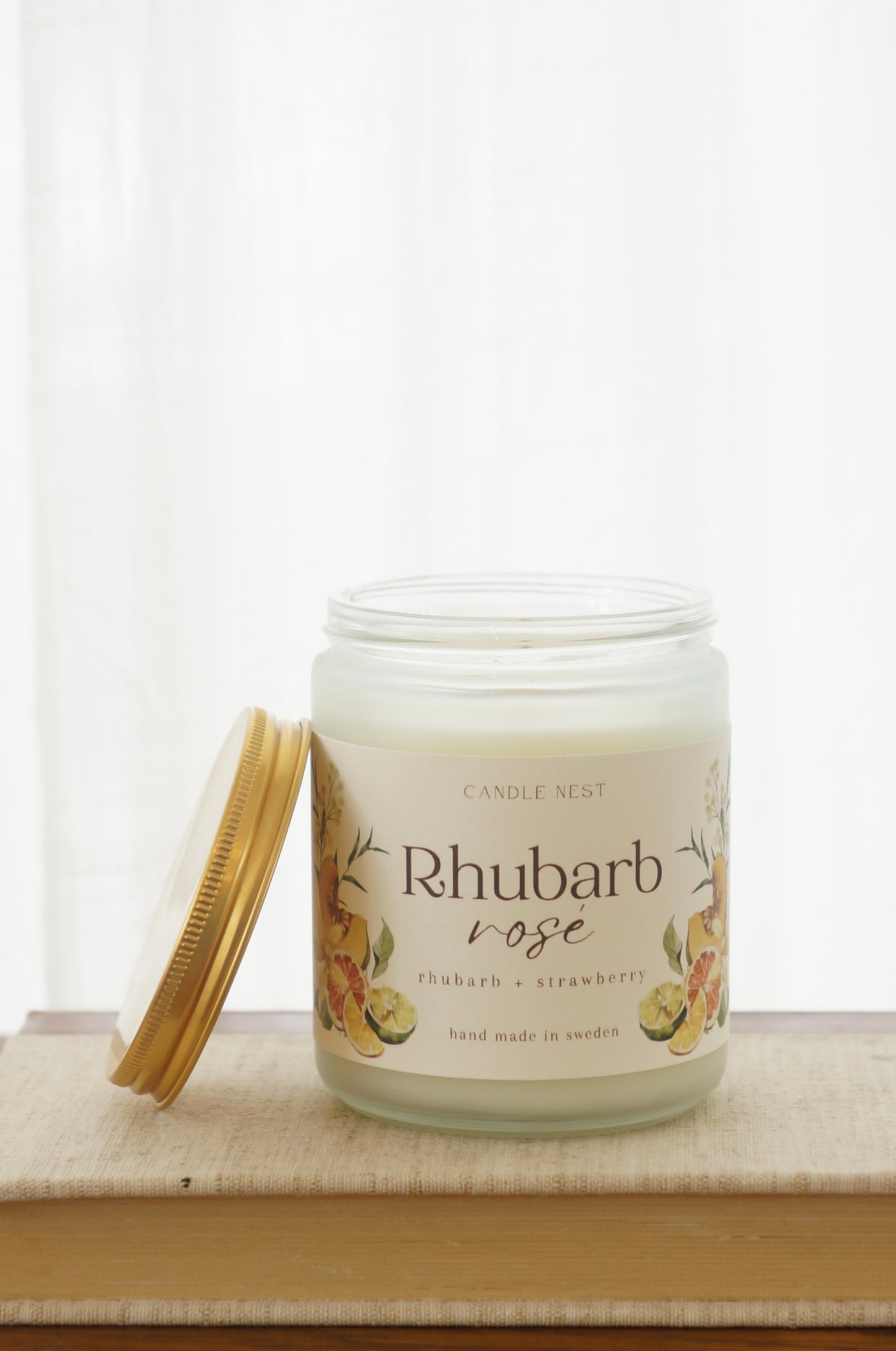 Ett doftljus i en frostad vit glasburk står på en bok. Runt ljuset finns ett off-white papper med bilder av frukter och texten "Rhubarb rosé". Mot ljuset står burken skruvlock i guld lutat. 