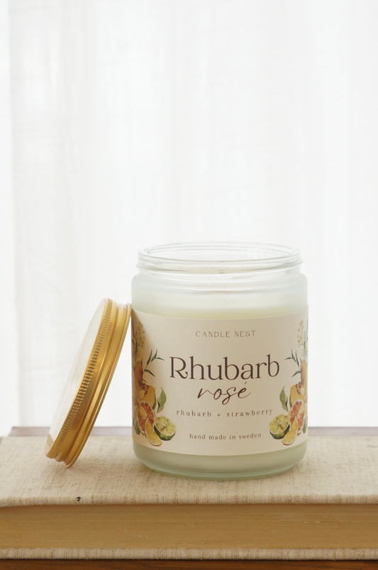 Ett doftljus i en frostad vit glasburk står på en bok. Runt ljuset finns ett off-white papper med bilder av frukter och texten "Rhubarb rosé". Mot ljuset står burken skruvlock i guld lutat. 