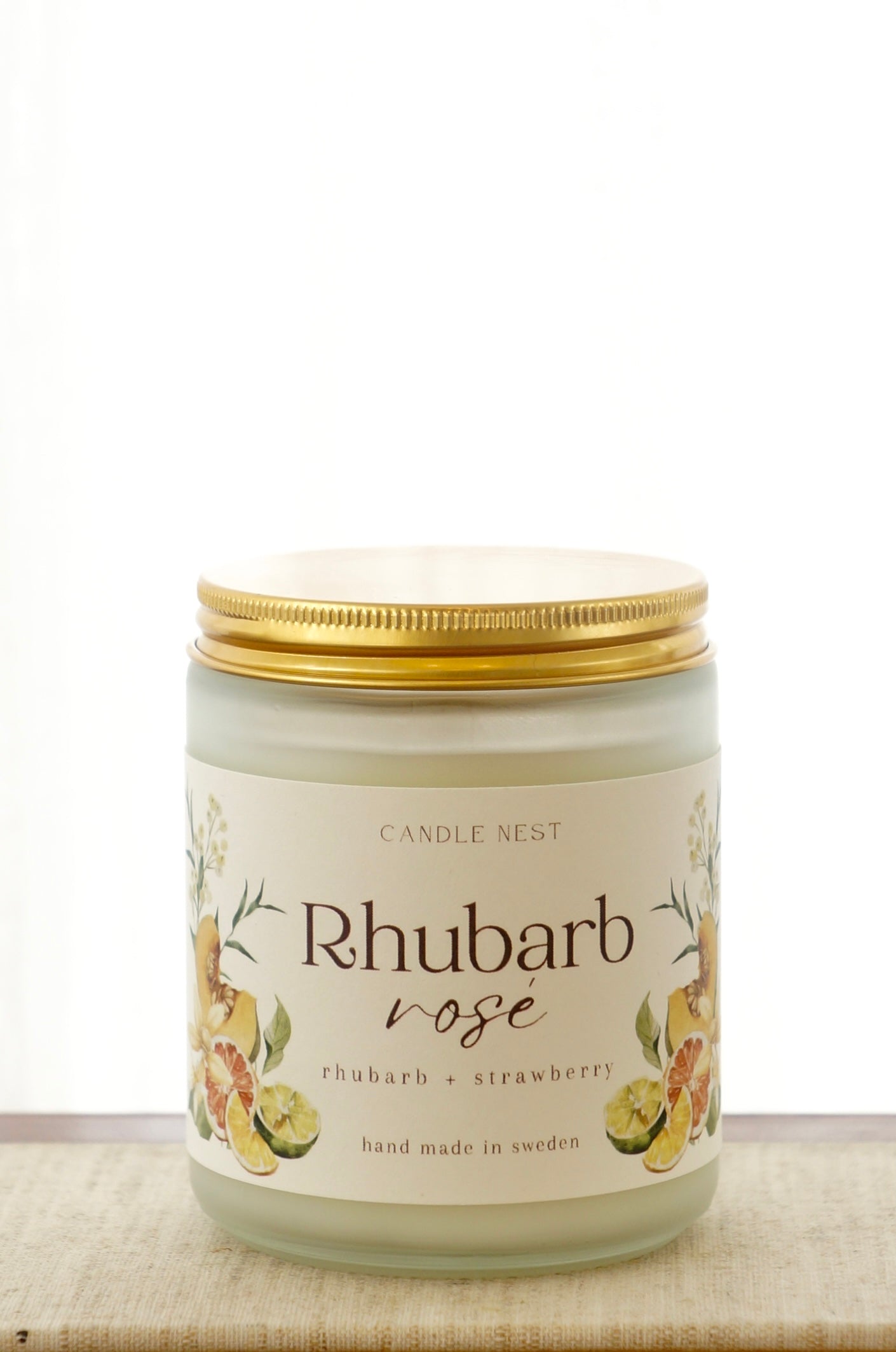 Ett doftljus i en frostad vit glasburk står på en bok. Runt ljuset finns ett off-white papper med bilder av frukter och texten "Rhubarb rosé". Burken är förslutet med ett skruvlock i guld.