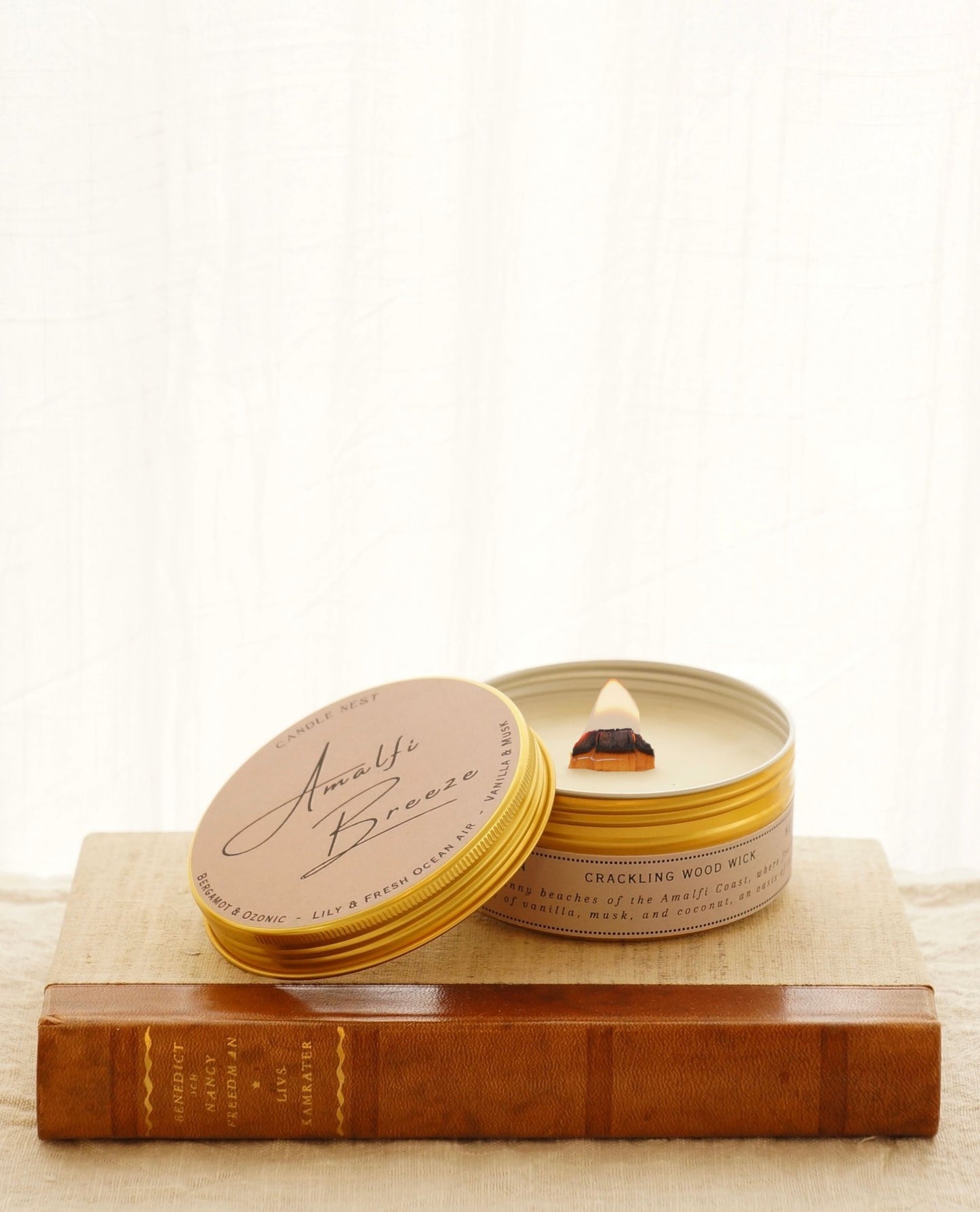 candle nest Dofftljus i en alluminiumburk i guld som står på en äldre bok med läderrygg. ljuset har en veke av trä som brinner. burkens runda lock står lutat mot burken och texten "amalfi breexe" står skrivet på locket med snikliga bokstäver.