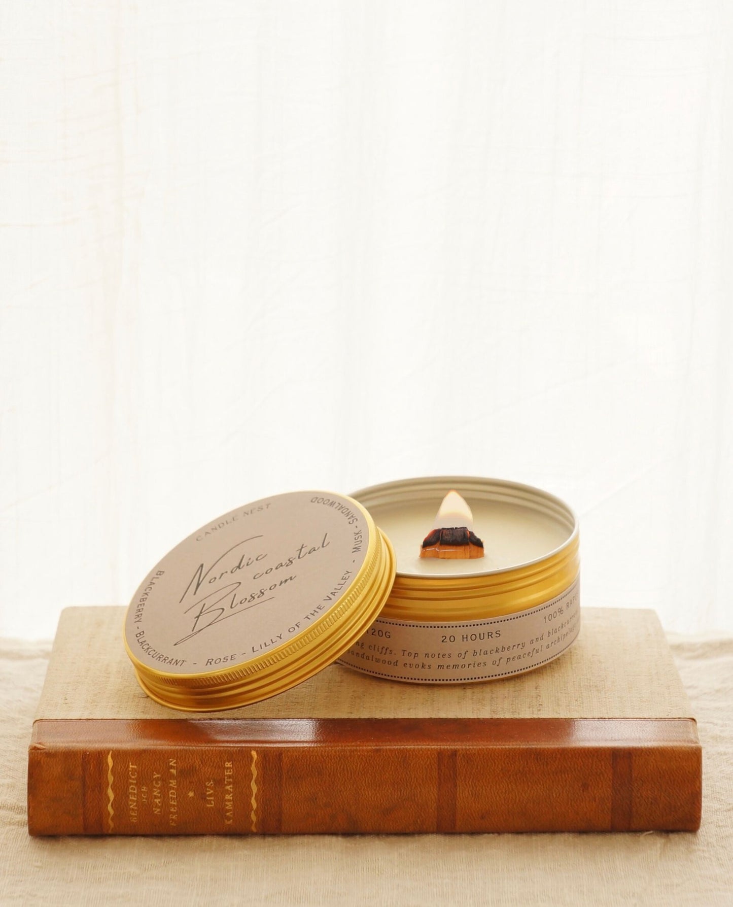Candle nest doftljus "Nordic coastal blossom" står med locket av på en bok med läderrygg. Ljuset kommer i en guldig metallburk. Locket ligger lutat mot sidan av burken så man ser vax och bred veke av trä som brinner med klar bred låga.