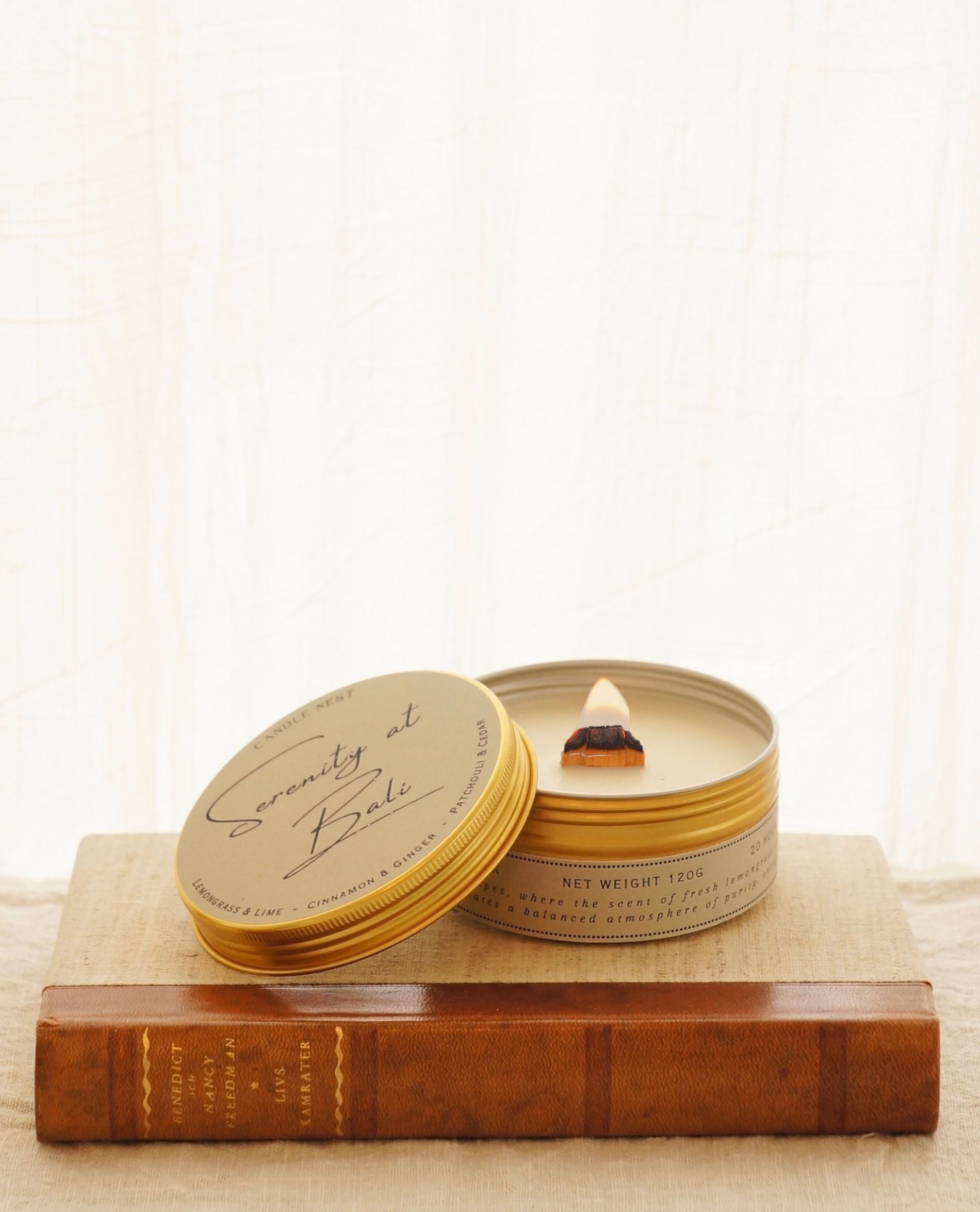 candle nest doftljus i en guldig rund burk av metall står på en äldre bok med läderrygg. Locket ligger lutat mot sidan av burken och exponerar vax och veke av trä som brinner med klar bred låga. på locket står det skrivet "Serenity at Bali" med snirklig font.