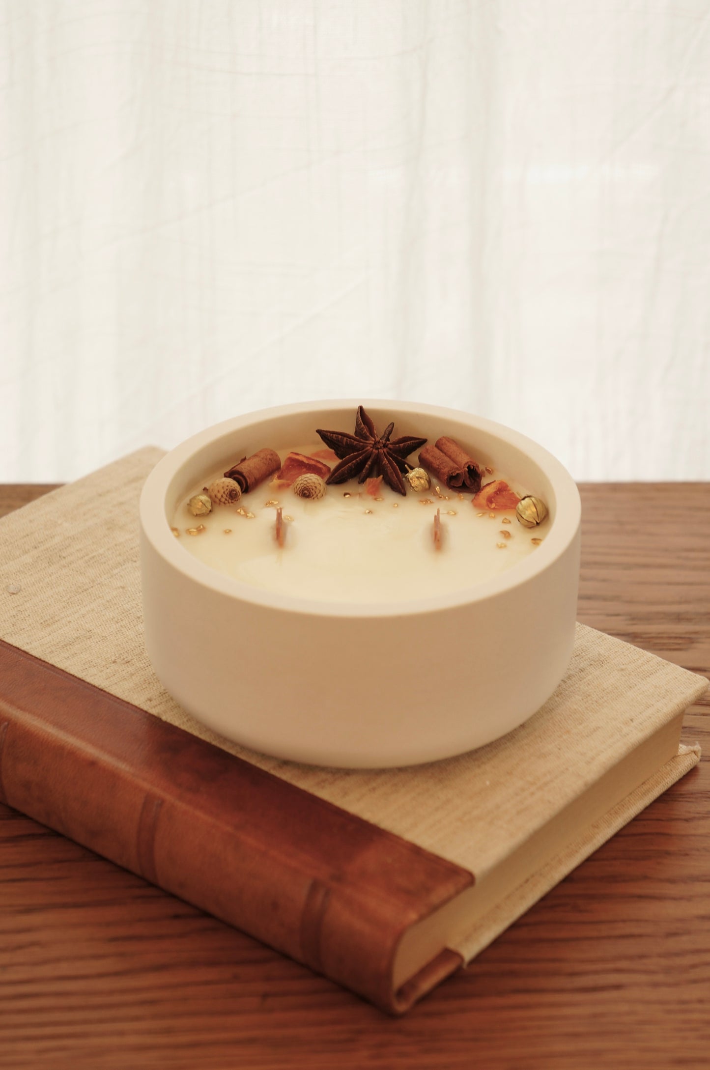 Doftljus i en vit gispskål står på en äldre bok med läderrygg. Ljusets ovansida är dekorerat med små bitar av clementin, kanlestänger och guldfärgade linfrökapsalar. Ljuset är släkt och de två vekarna av trä syns. 