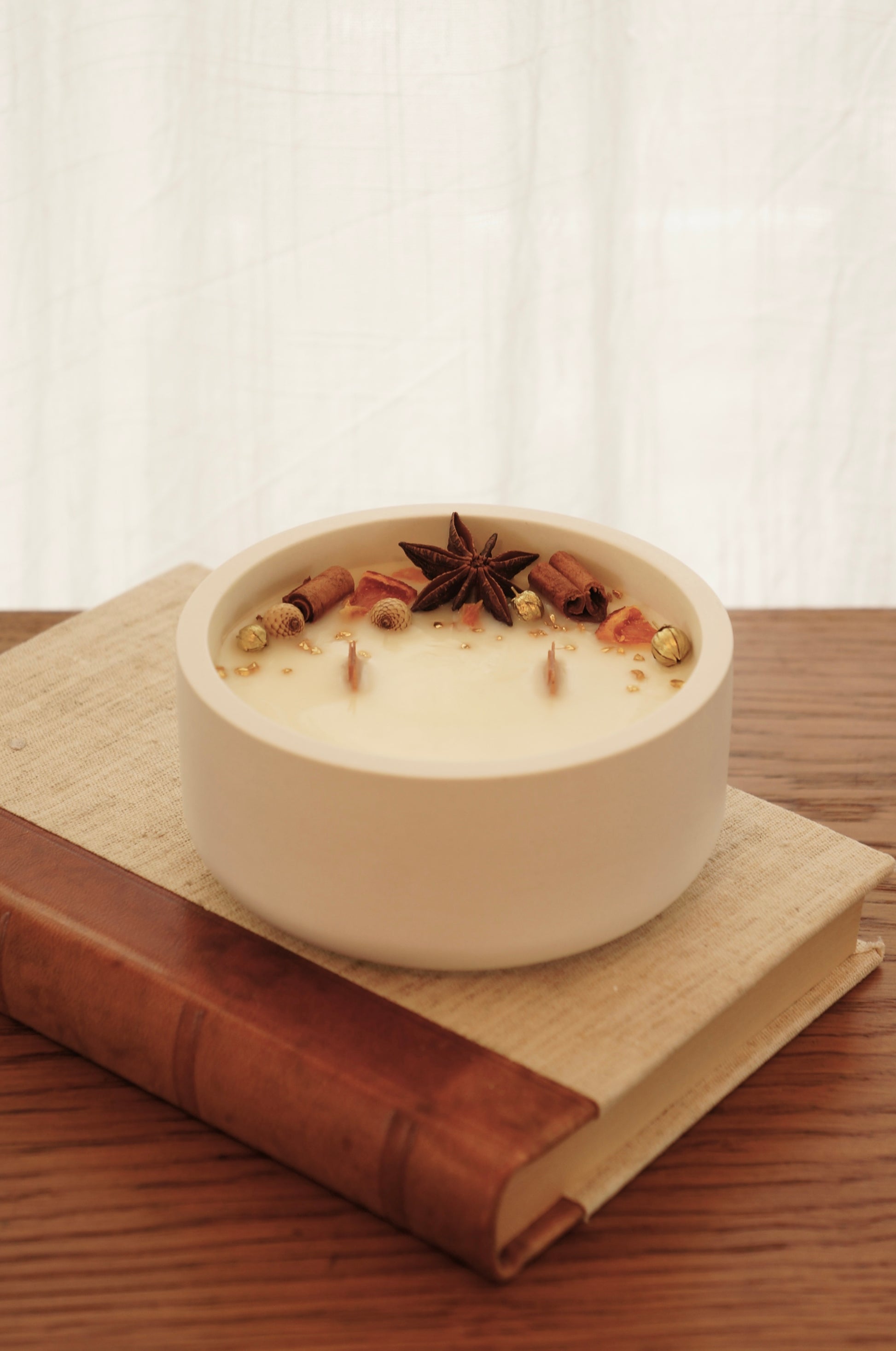 Doftljus i en vit gispskål står på en äldre bok med läderrygg. Ljusets ovansida är dekorerat med små bitar av clementin, kanlestänger och guldfärgade linfrökapsalar. Ljuset är släkt och de två vekarna av trä syns. 