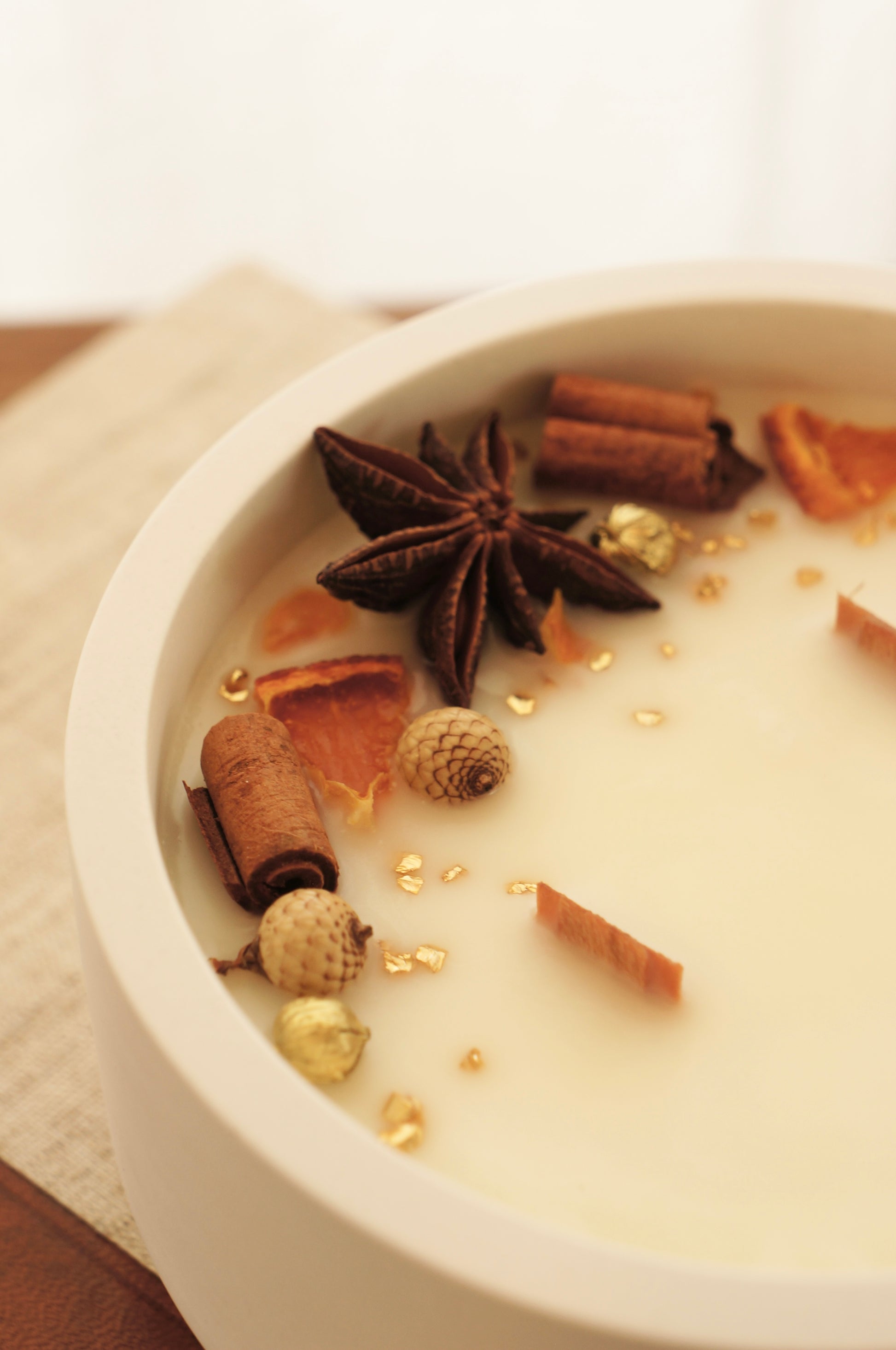 Närbild på ett Doftljus i en vit gispskål.  Ljusets ovansida är dekorerat med små bitar av clementin, kanlestänger och guldfärgade linfrökapsalar.