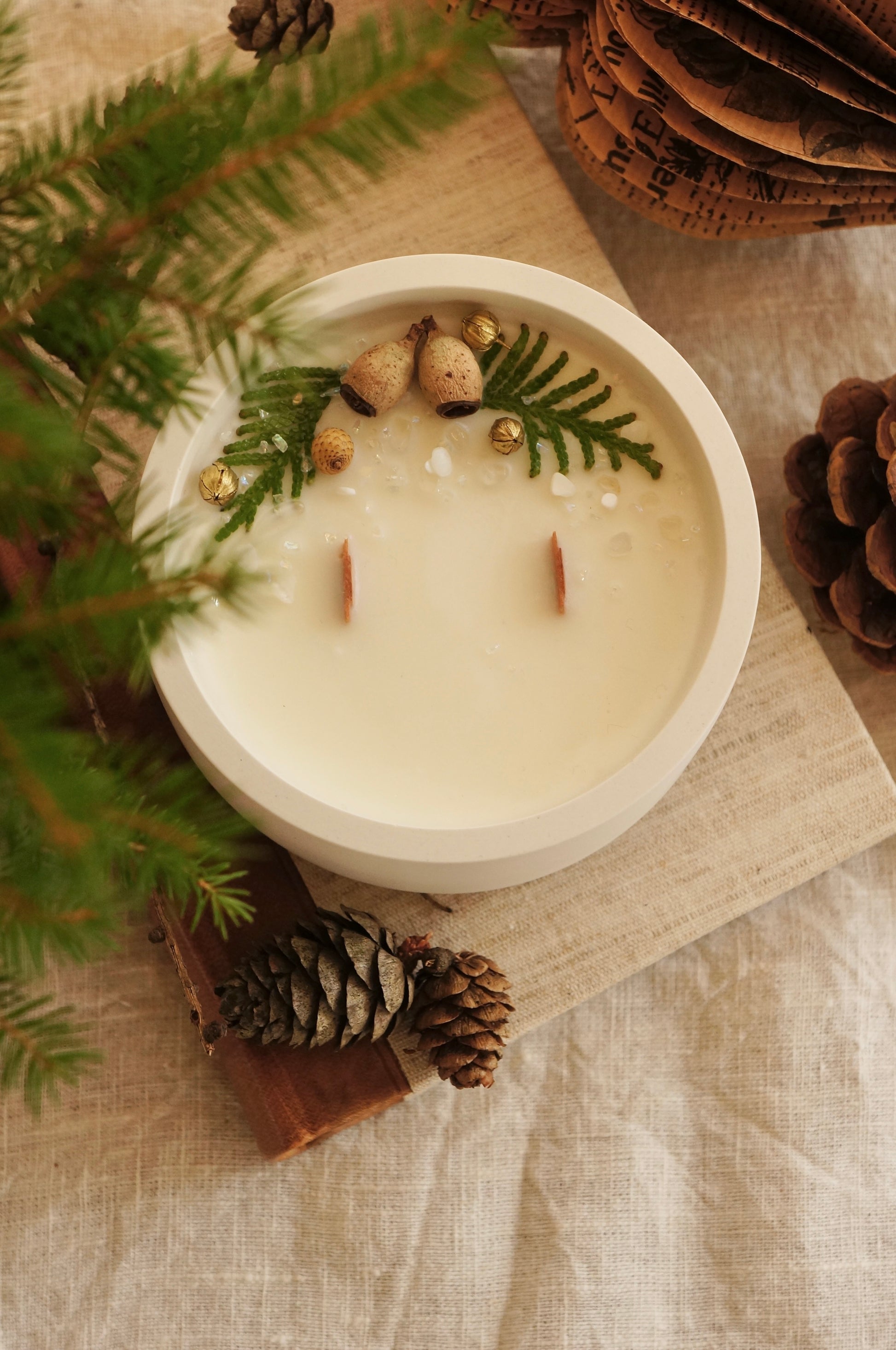 Ett doftljus i en vit gipsskål står på en äldre bok med läderrygg och framsida av linnetyg. Ljuset har två bredare vekar av trä och ovansidan är dekorerad med kvistar av thuja, små kottar och guldfärgade linfrökapslar. I förgrunden och bakgrunden syns kottar och granris.