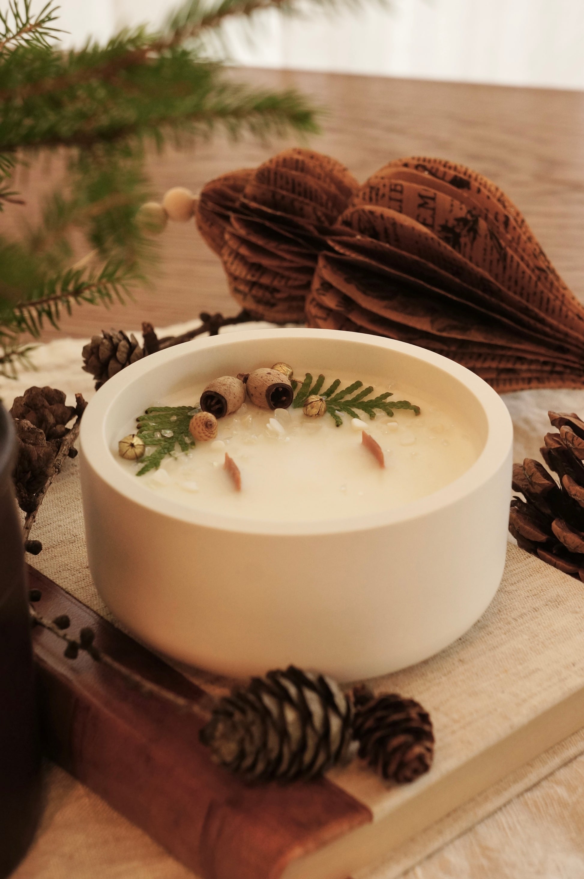 Ett doftljus i en vit gipsskål står på en äldre bok med läderrygg och framsida av linnetyg. Ljuset har två bredare vekar av trä och ovansidan är dekorerad med kvistar av thuja, små kottar och guldfärgade linfrökapslar. I förgrunden och bakgrunden syns kottar och granris.