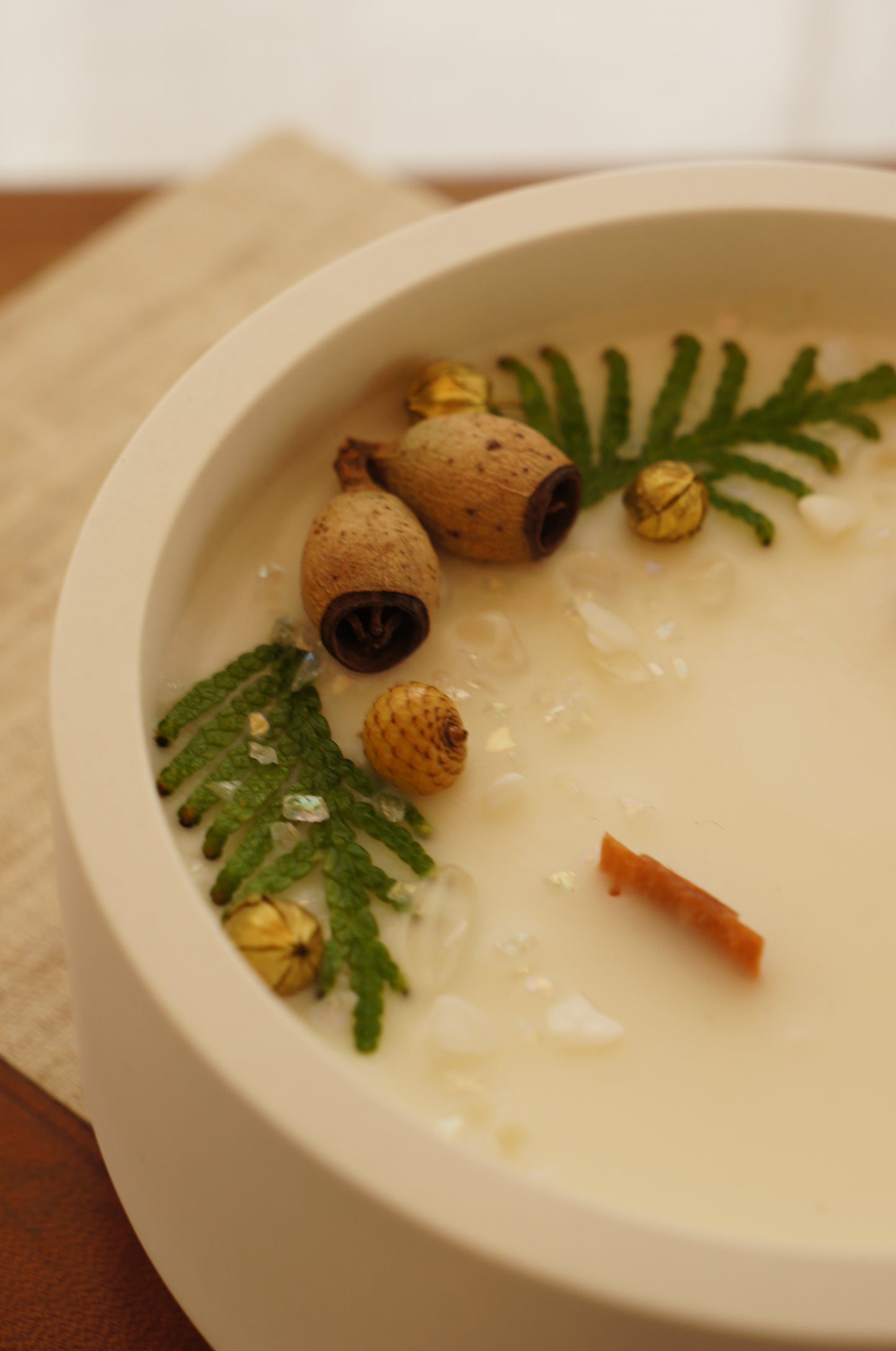 Ett doftljus i en vit gipsskål. Ljuset har två bredare vekar av trä och ovansidan är dekorerad med kvistar av thuja, små kottar och guldfärgade linfrökapslar. 