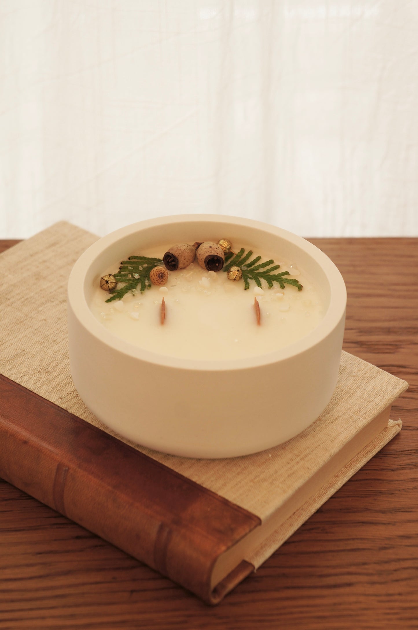 Ett doftljus i en vit gipsskål står på en äldre bok med läderrygg och framsida av linnetyg. Ljuset har två bredare vekar av trä och ovansidan är dekorerad med kvistar av thuja, små kottar och guldfärgade linfrökapslar.