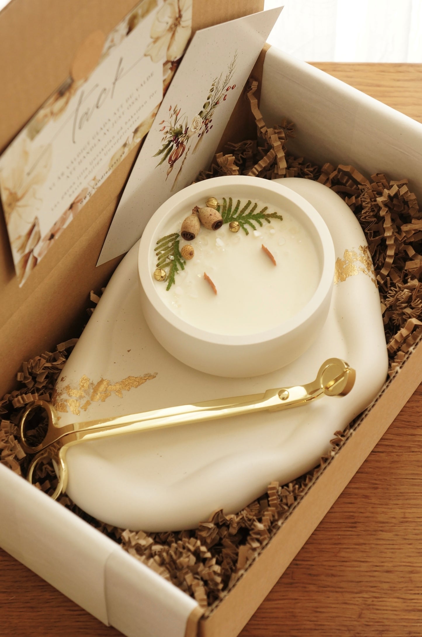 en presentlåda i papp står på et bord, i lådan finns ett molnformat vitt ljusfat med dekor i guld. på fatet står ett doftljus i en vit gipsskål. Ljuset har två vekar av trä. Ljusets ovansida är dekorerat med små kvistar av thuja och minikottar. En vektång i guld ligger på fatet i lådan.