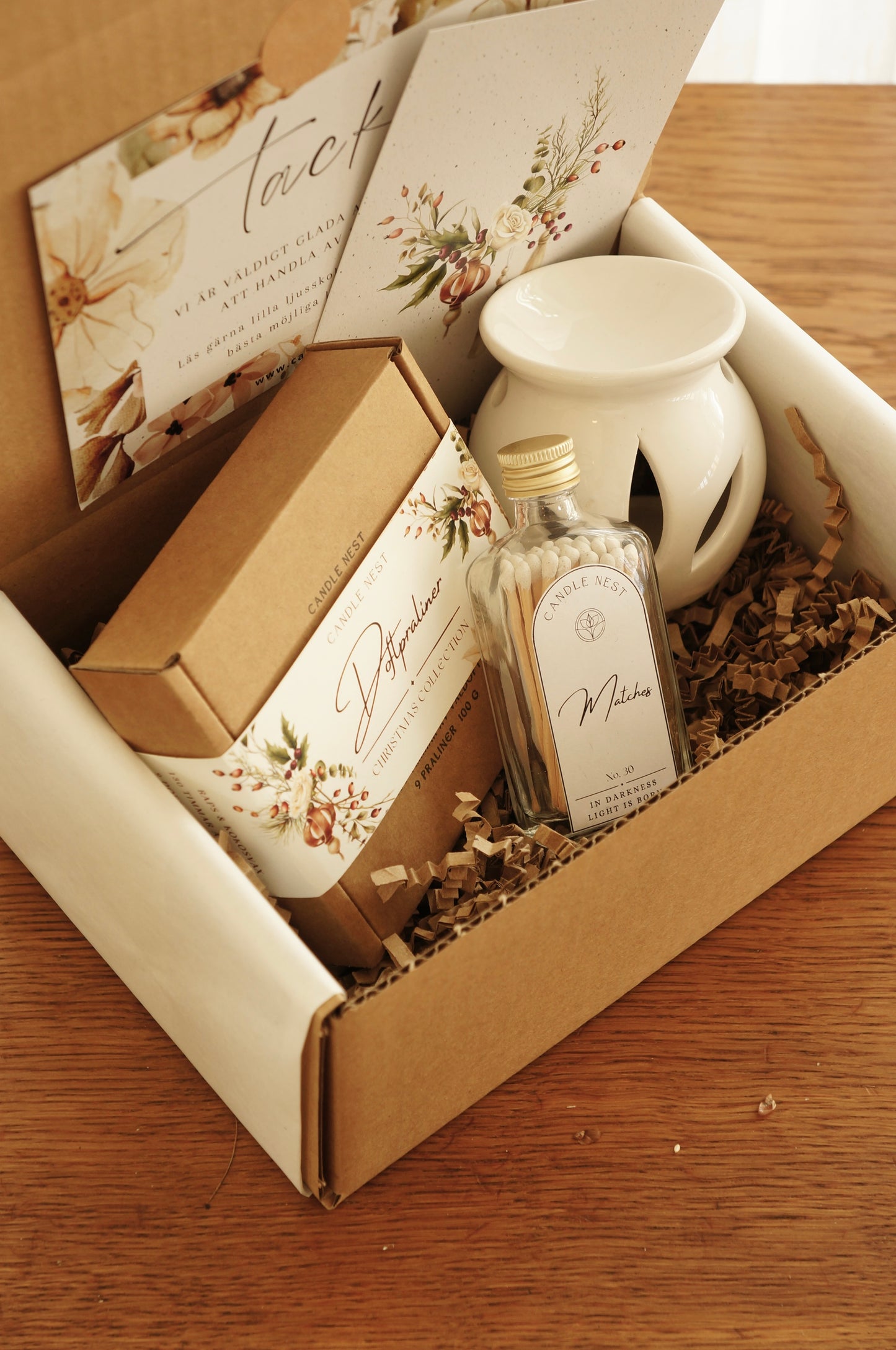 En låda i papp står på ett bord. i lådan ligger en ask med julens doftpraliner, en vit aromalampa, en flaska med tändstickor.