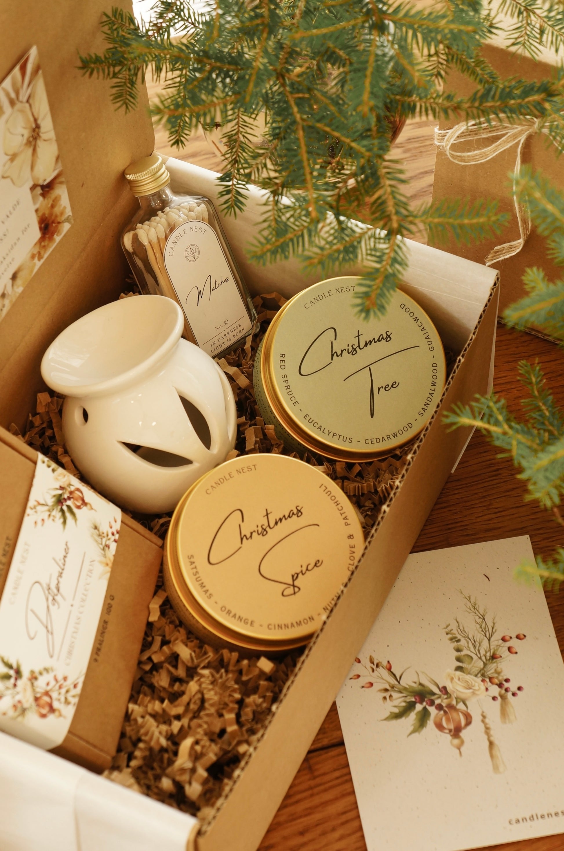 En låda i papp står på ett bord. i lådan ligger en ask med julens doftpraliner, en vit aromalampa, en flaska med tändstickor samt två travel candles i dofterna Christmas spice och Christmas Tree