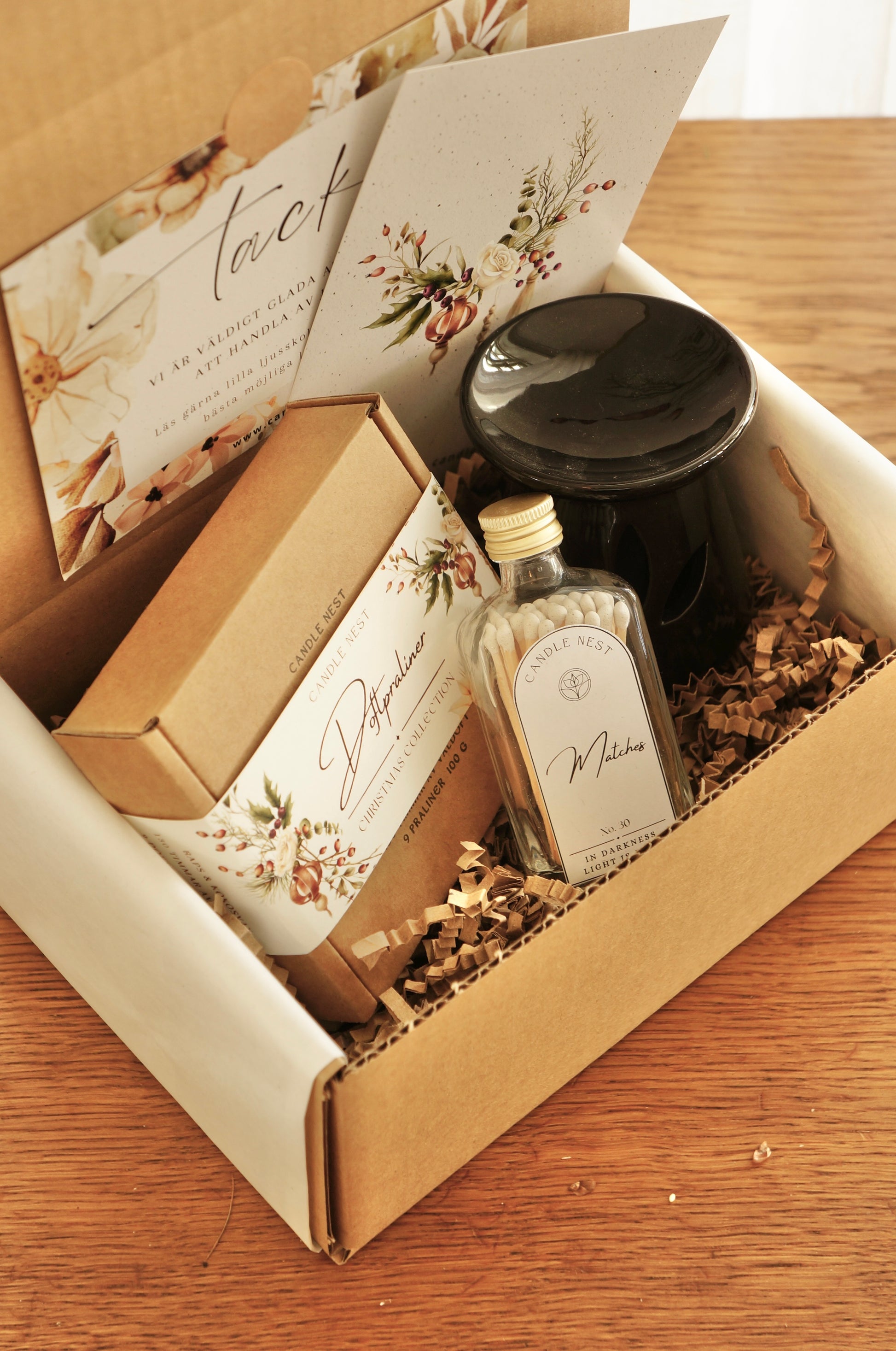 En låda i papp står på ett bord. i lådan ligger en ask med julens doftpraliner, en svart aromalampa, en flaska med tändstickor.