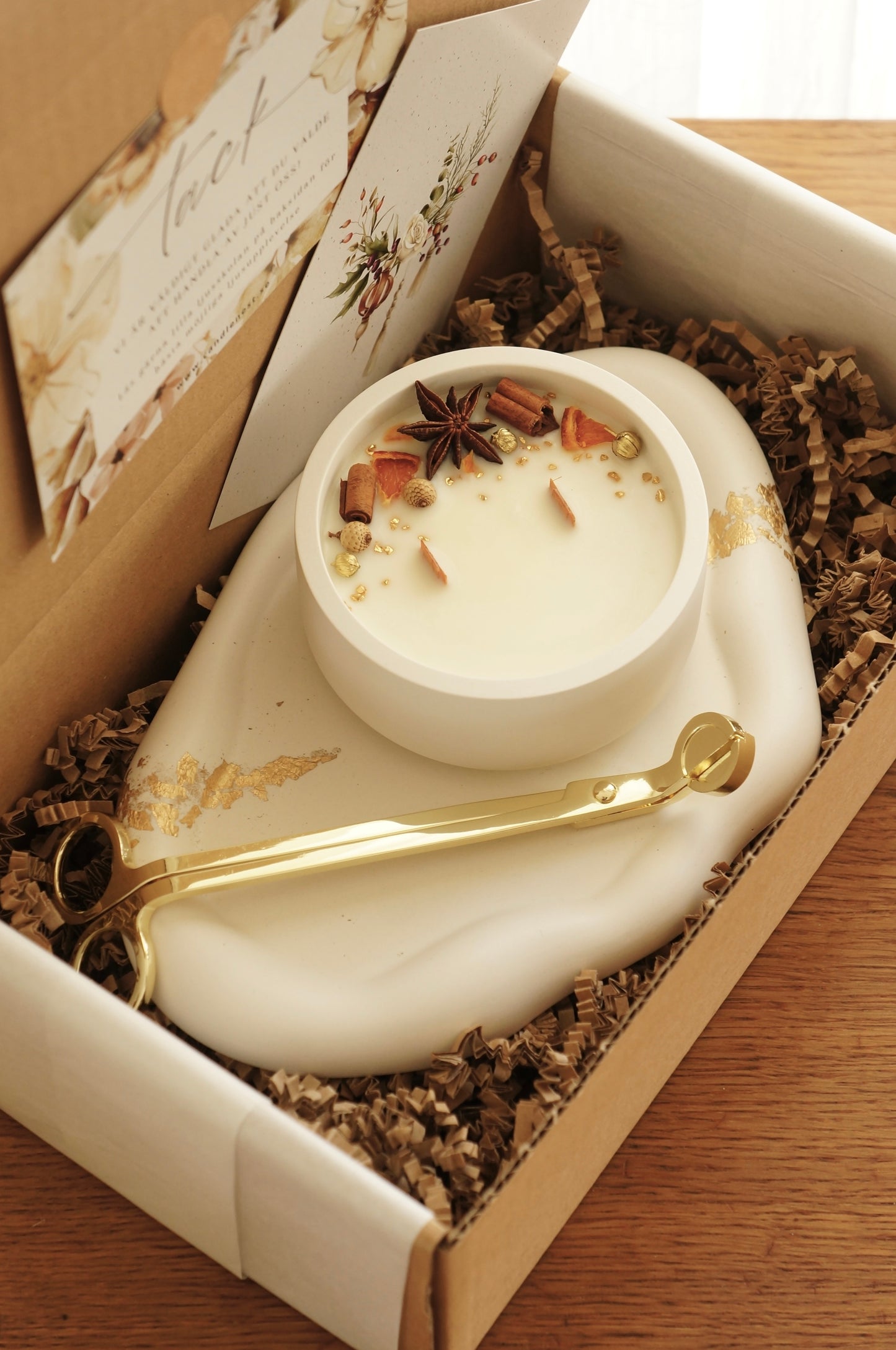 en presentlåda i papp står på et bord, i lådan finns ett molnformat vitt ljusfat med dekor i guld. på fatet står ett doftljus i en vit gipsskål. Ljuset har två vekar av trä. Ljusets ovansida är dekorerat med små bitar av clementin, stjärnanis och kanelstång. En vektång i guld ligger på fatet i lådan.