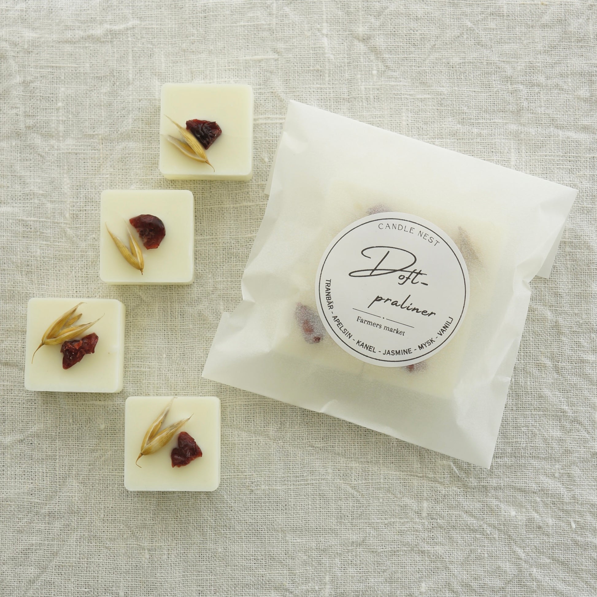 4 st doftpraliner av doften "Farmers market" och en påse med doftpraliner ligger på en vit linneduk. Doftvaxen är dekorerade med torkad havre och tranbär.