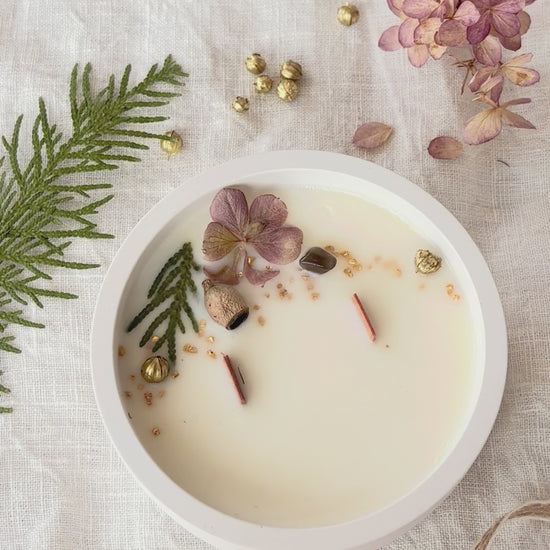 Videon visar ett runt doftljus uppifrån. Ljuset är dekorerat med torkade växter. En pincet lägger sakta dit ett blad. 