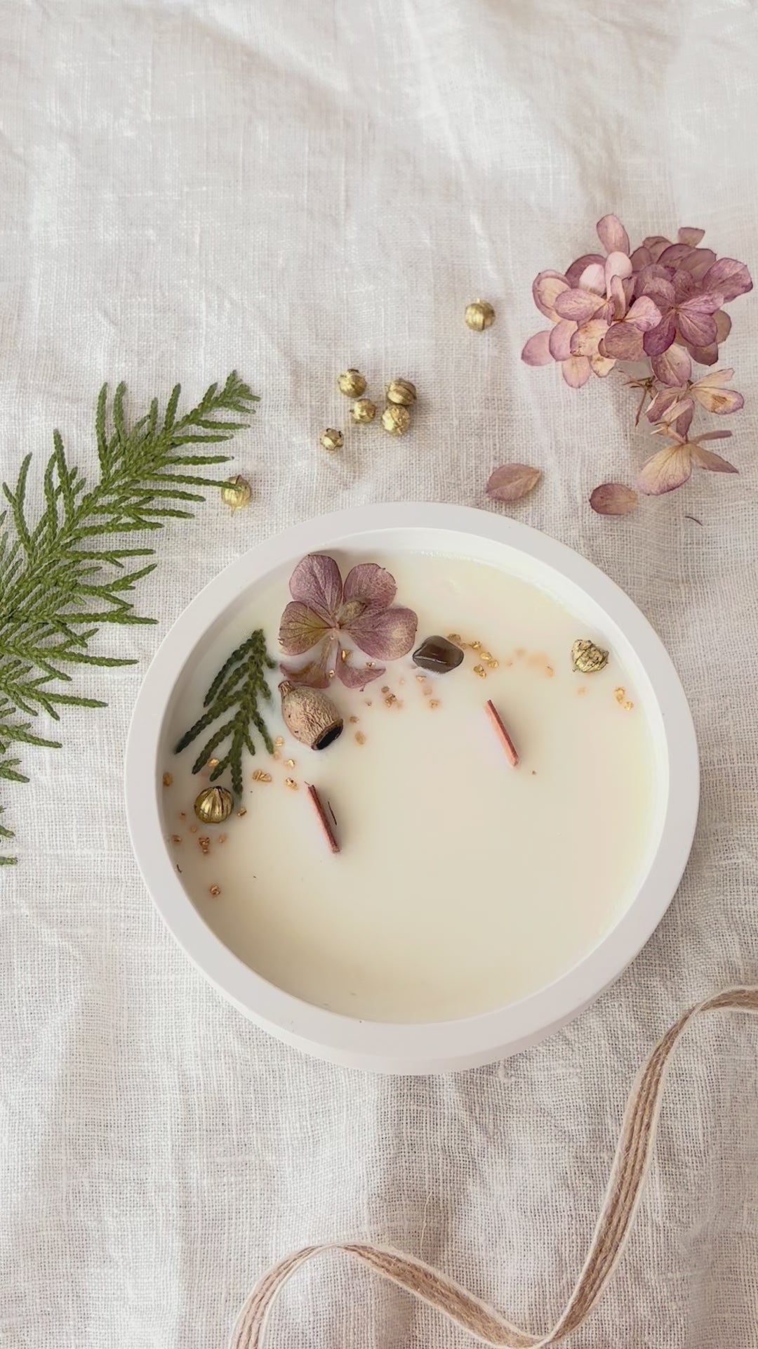 Videon visar ett runt doftljus uppifrån. Ljuset är dekorerat med torkade växter. En pincet lägger sakta dit ett blad. 